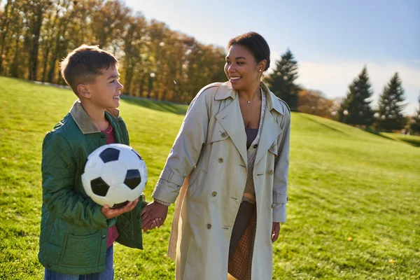 Niño afroamericano feliz sosteniendo pelota de fútbol, caminando con mamá en el campo verde, ropa de abrigo, otoño - foto de stock