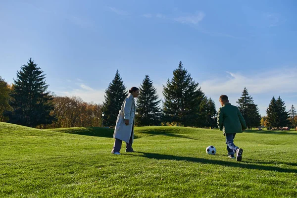 Alegre afroamericano chico jugando fútbol con feliz madre en verde campo, fútbol, otoño, correr - foto de stock