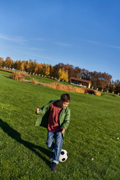 Chico afroamericano deportivo en ropa de abrigo otoñal jugando fútbol en el campo verde, fútbol, otoño - foto de stock