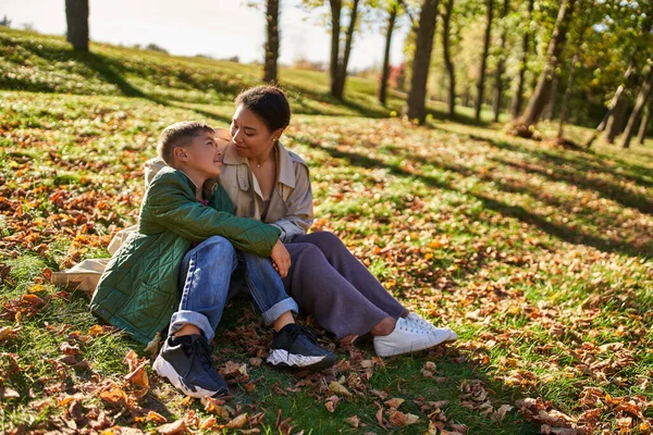 Amor maternal, alegre mujer afroamericana e hijo sentado en la hierba con hojas doradas, otoño - foto de stock