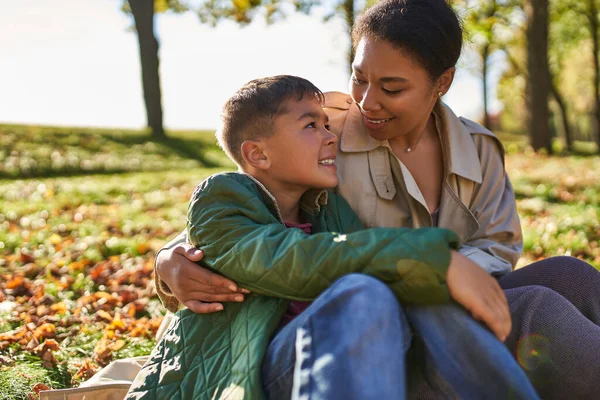 Motherly love, cheerful african american woman and son hugging and sitting near golden leaves, fall — Stock Photo