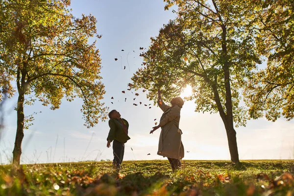 Silueta de la madre y el niño lanzando hojas de otoño, parque, temporada de otoño, divertirse, libertad - foto de stock