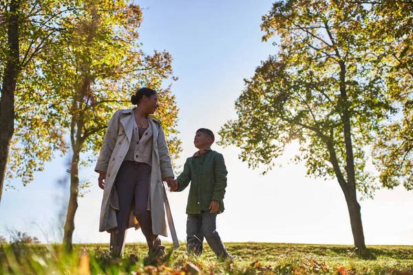 Madre y el niño afroamericanos tomados de la mano y caminando cerca de los árboles en el parque de otoño, temporada de otoño - foto de stock
