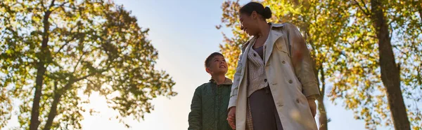 Madre e bambino afroamericani in capispalla che si tengono per mano vicino agli alberi nel parco autunnale, autunno, banner — Foto stock