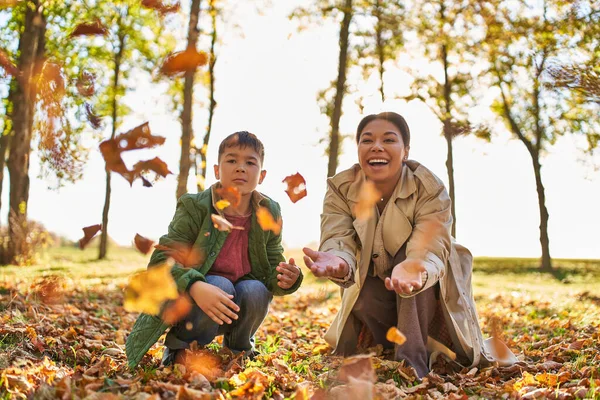 Recuerdos felices, madre e hijo gozosos lanzando hojas otoñales, temporada de otoño, familia afroamericana - foto de stock
