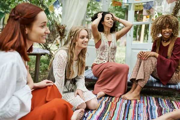 Mujeres alegres y multiétnicas en ropa de estilo boho hablando al aire libre en el centro de retiro - foto de stock