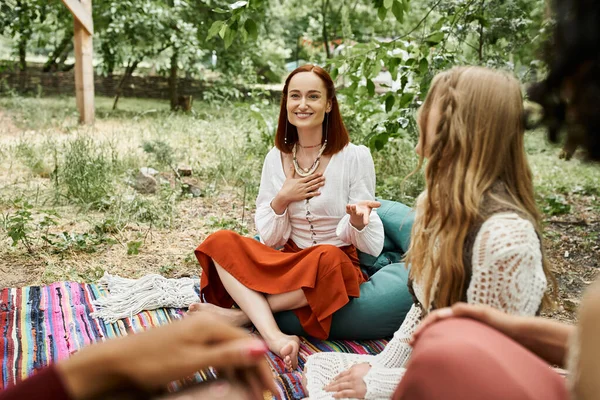 Smiling redhead woman in boho styled clothes talking to girlfriends on lawn in retreat center — Stock Photo