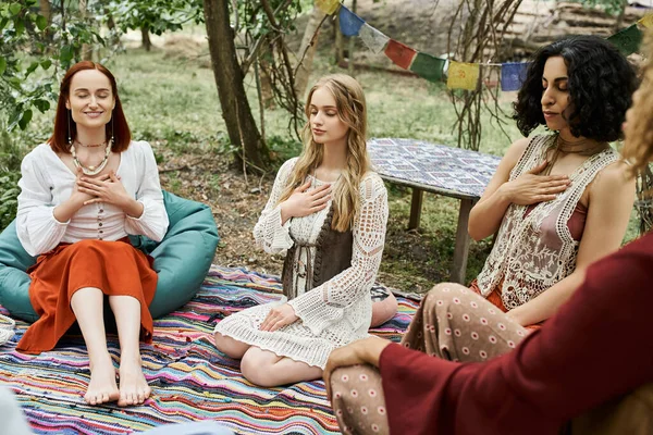 Young woman meditating near smiling and multiethnic girlfriends on meadow in retreat center — Stock Photo