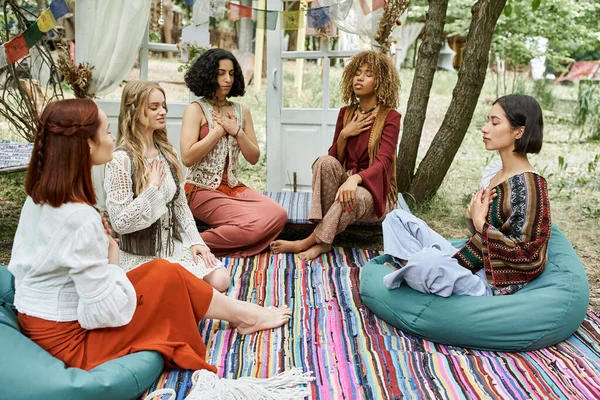 Mulheres elegantes e inter-raciais em roupas estilo boho meditando juntos no gramado no centro de retiro — Fotografia de Stock