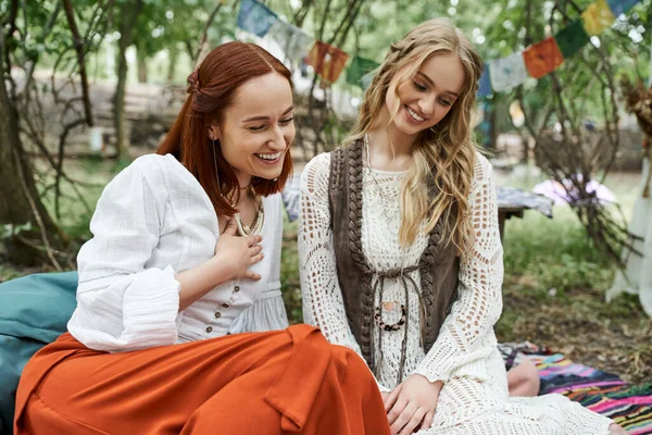 Laughing woman in boho styled clothes sitting near friend on carpet on lawn in retreat center — Stock Photo