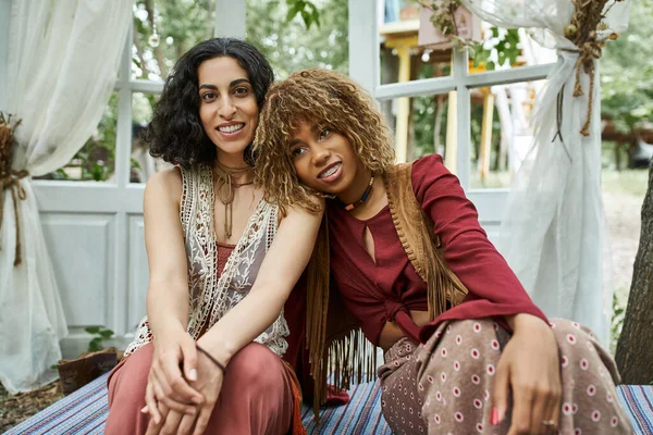 Mujeres multiétnicas sonrientes y de moda en ropa de estilo boho que pasan tiempo en el patio en el centro de retiro - foto de stock