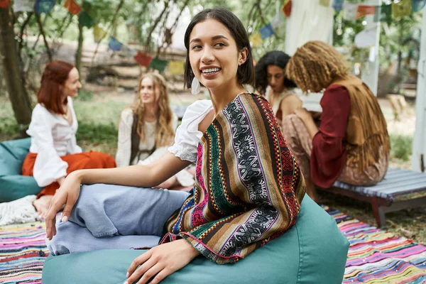 Молодая улыбающаяся женщина сидит на бобовом мешке рядом с размытыми многонациональными друзьями в центре отдыха — стоковое фото