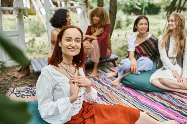 Smiling redhead woman looking at camera near blurred multiethnic friends outdoors in retreat center — Stock Photo