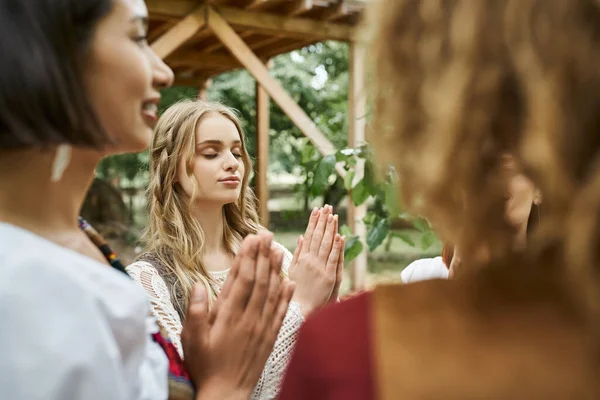 Giovane donna elegante in abito boho pregare vicino amici sorridenti all'aperto nel centro di ritiro — Foto stock