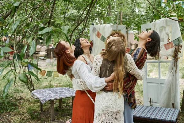 Mujeres alegres e interracial amigas en trajes boho abrazándose al aire libre en el centro de retiros - foto de stock