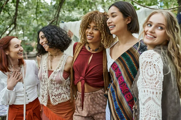 Mujeres multiétnicas positivas y de moda en ropa boho abrazándose al aire libre en el centro de retiros - foto de stock