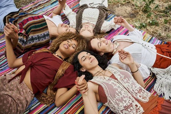 Високий кут зору усміхнених багатоетнічних жінок, що тримаються за руки і лежать на ковдрі в центрі відступу — стокове фото
