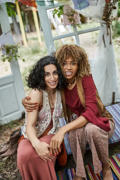 Smiling interracial girlfriends in boho outfits hugging and looking at camera in retreat center — Stock Photo