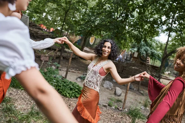 Positive multiethnic girlfriends holding hands and dancing on meadow in retreat center — Stock Photo