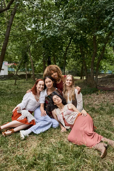 Fröhliche und stilvolle interrassische Freundinnen, die sich umarmen, während sie auf dem Rasen im Rückzugszentrum sitzen — Stockfoto