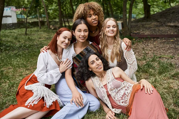 Trendy and smiling interracial girlfriends hugging while spending time on lawn in retreat center — Stock Photo