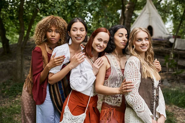 Trendy and carefree interracial girlfriends in boho outfits hugging outdoors in retreat center — Stock Photo