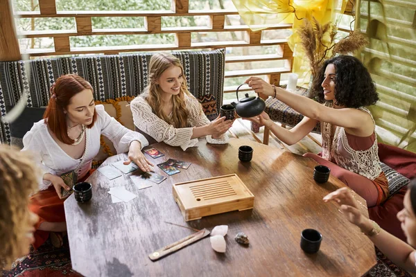 Multiracial woman pouring tea near girlfriends reading tarot cards in retreat center — Stock Photo