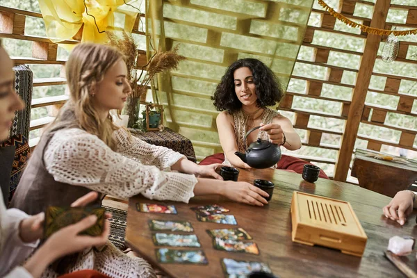 Stilvolle multirassische Frau schenkt Tee ein, während junge Freundinnen im Ferienhaus Tarot-Karten lesen — Stockfoto