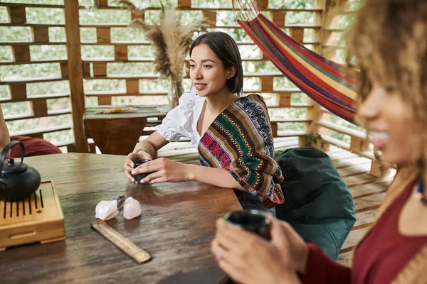 Happy young woman in boho style clothes drinking tea at wooden table in retreat center — Stock Photo