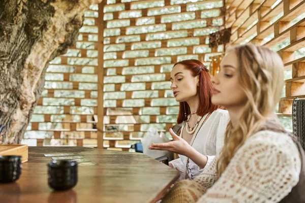 Woman pointing at tarot cards near blurred girlfriend in cottage on retreat center — Stock Photo