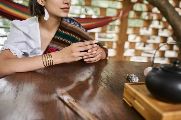 Vista recortada de la mujer joven en ropa de estilo boho sentado con taza de té en la mesa de madera - foto de stock