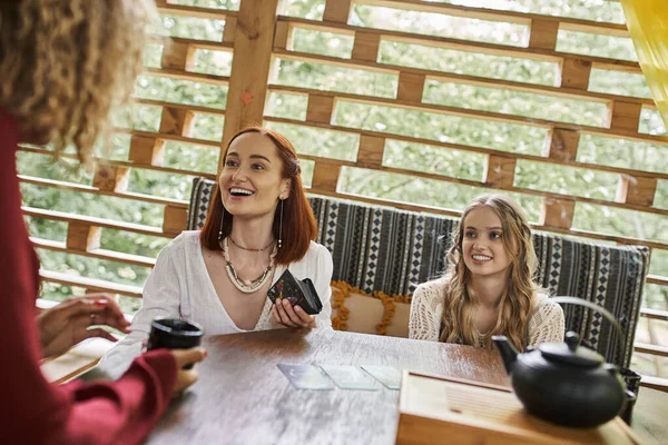 Cheerful woman holding tarot cards near multiethnic girlfriends at wooden table in retreat center — Stock Photo