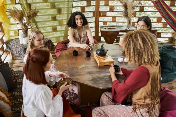Mujeres multiétnicas felices hablando en la mesa de madera cerca de tetera y tazas en casa de campo del centro de retiro - foto de stock