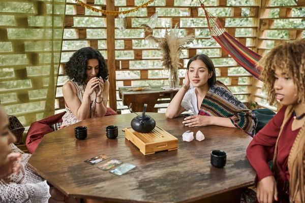 Multiracial woman drinking tea near girlfriends and tarot cards on wooden table, women retreat — Stock Photo