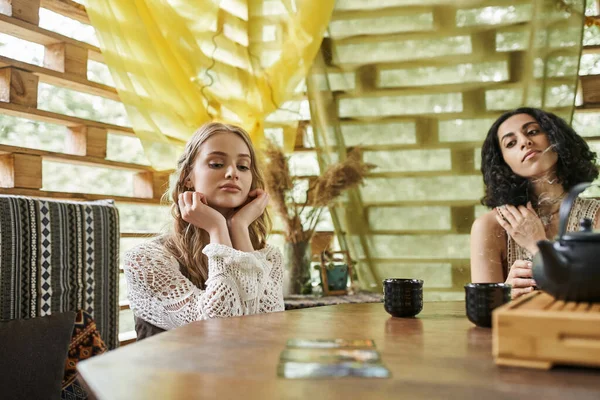 Mujer joven y soñadora cerca de la novia multirracial y tazas de té en la mesa de madera en acogedora casa de campo - foto de stock