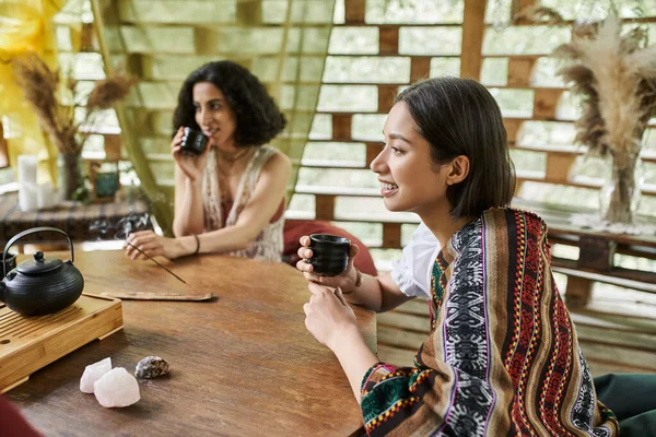 Cheerful young woman with tea cup near multiracial girlfriend at wooden table in retreat center — Stock Photo