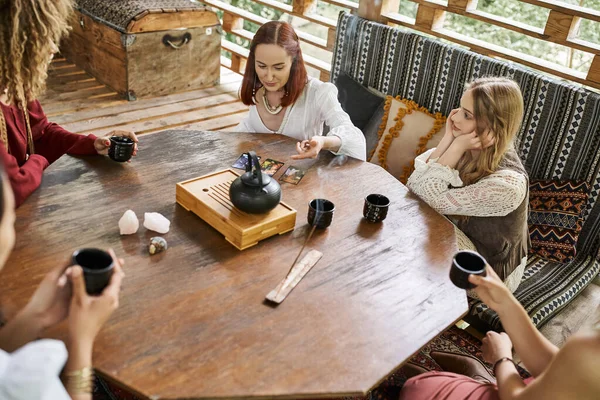 Woman reading tarot cards near multiethnic girlfriends and tea on wooden table in cozy house — Stock Photo