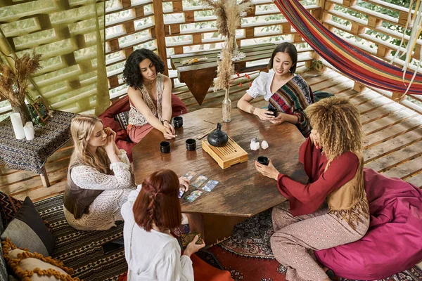 Multiethnic boho style girlfriends at wooden table with tarot cards and tea in retreat center — Stock Photo