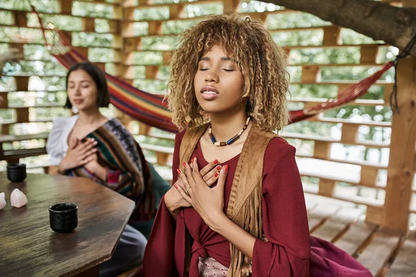 Afroamerikanerin in Boho-Klamotten meditiert mit geschlossenen Augen in der Nähe einer verschwommenen Freundin — Stockfoto
