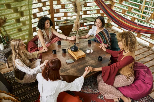 Sorrindo namoradas multirraciais de mãos dadas e meditando à mesa de madeira com xícaras de chá — Fotografia de Stock