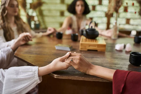Cropped view of multiethnic girlfriends holding hands while meditating in retreat center — Stock Photo