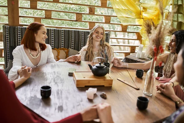 Joyeuses copines se tenant la main et méditant près de théière et tasses sur table en bois dans le chalet — Photo de stock