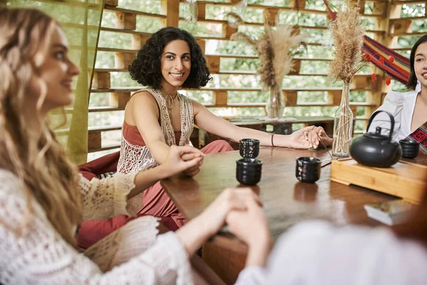 Happy multiracial woman holding hands and meditating with girlfriends near tea on wooden table — Stock Photo