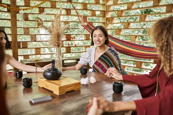 Mujer feliz en ropa de estilo boho meditando con amigas en la mesa con té en el centro de retiro - foto de stock