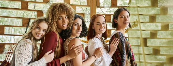 Joyful, stylish multiethnic girlfriends smiling at camera and embracing in retreat center, banner — Stock Photo