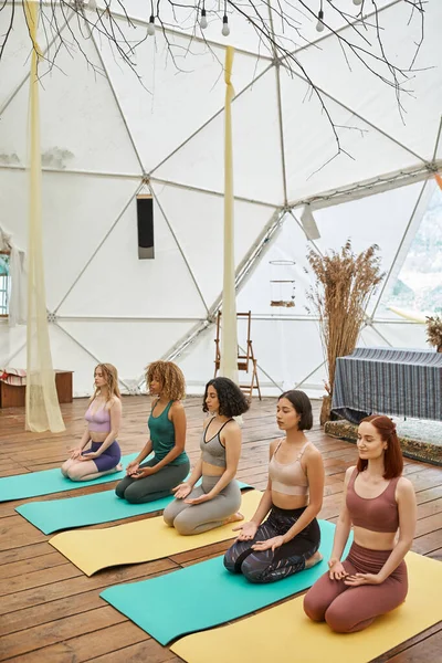 Multiracial girlfriends in sportswear meditating in thunderbolt pose in modern retreat center — Stock Photo