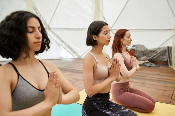 Multiracial woman with closed eyes and praying hands meditating near girlfriends in retreat center — Stock Photo