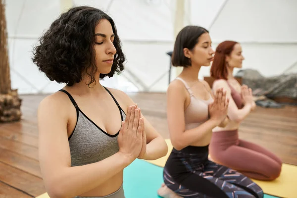 Mulher multirracial com olhos fechados e mãos de oração praticando ioga com namoradas, harmonia — Fotografia de Stock