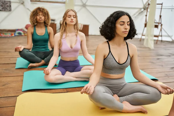 Mujeres multiétnicas en ropa deportiva sentadas en colchonetas de yoga y meditando en pose de loto, estilo de vida saludable - foto de stock
