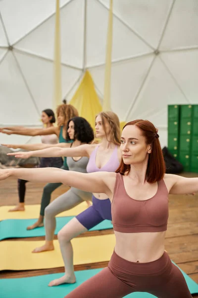 Femme regardant loin tout en pratiquant le yoga dans la pose guerrière près de copines multiethniques — Photo de stock